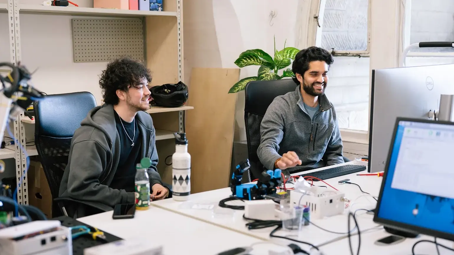 Two men sitting at desks in an office, one working on a computer and the other watching his screen
