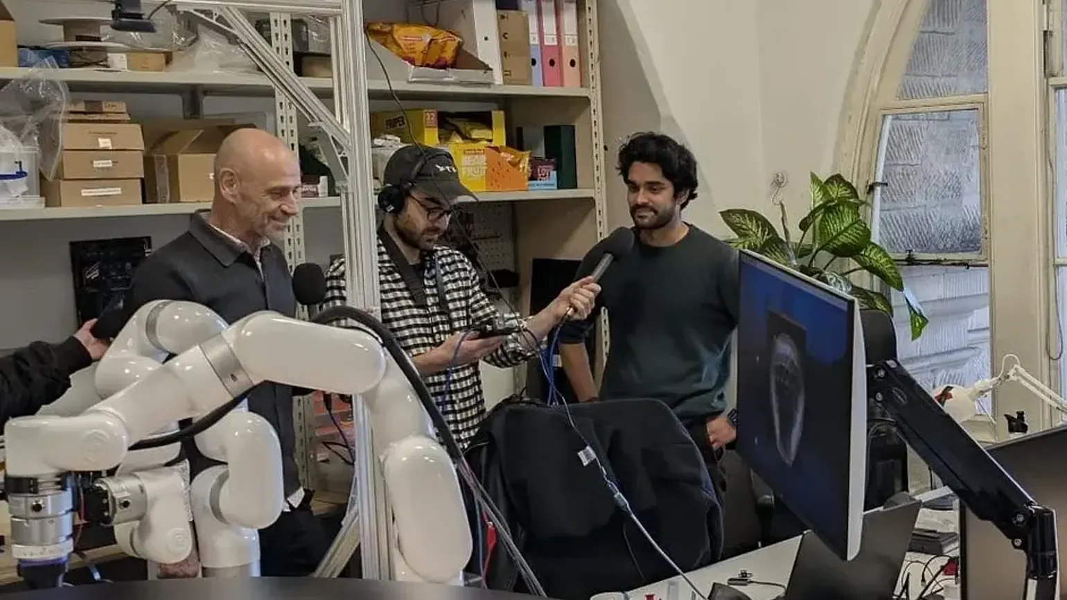 Three men in an office, one man holding a microphone in front of another who is being interviewed.