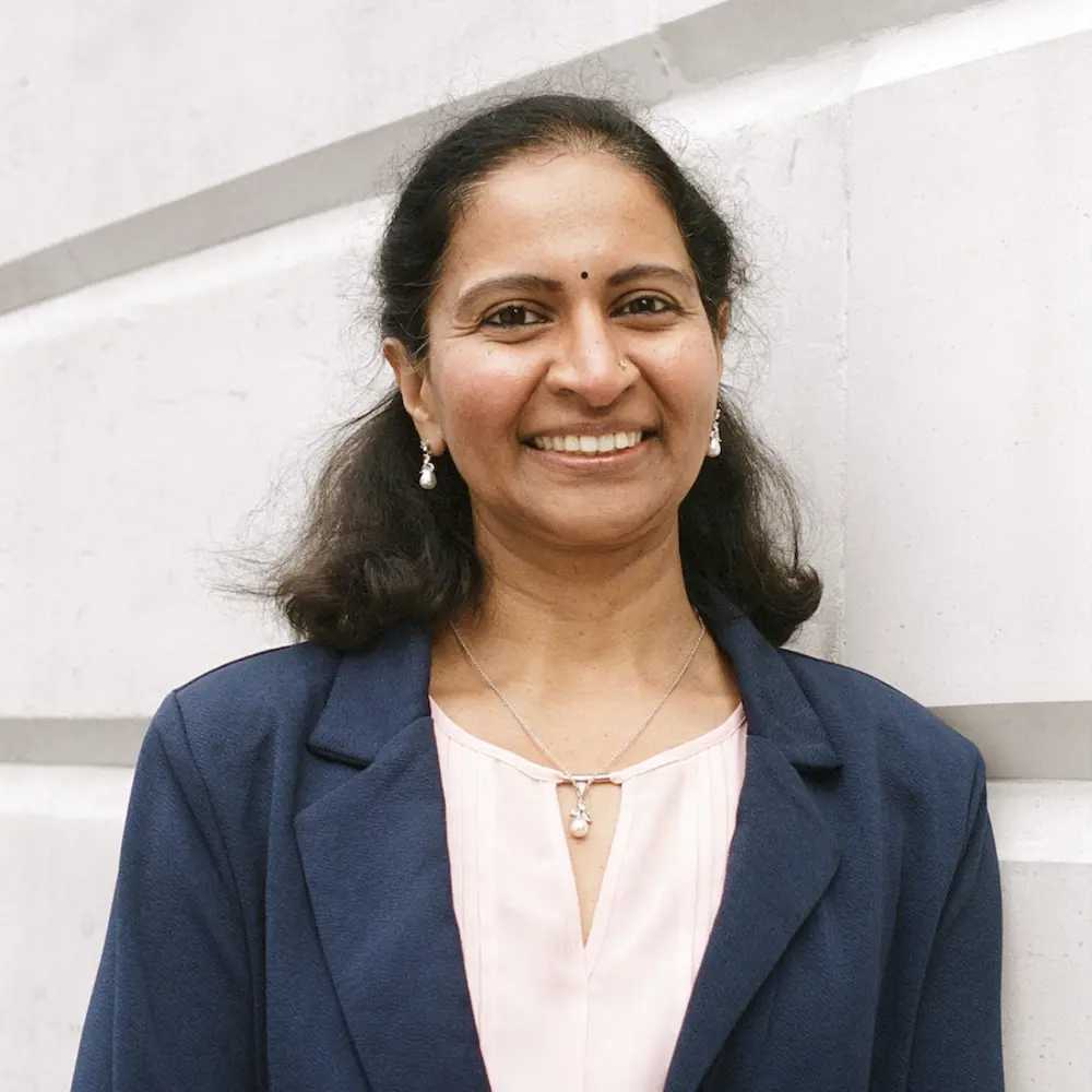 A photo of Radhika Gudipati smiling and standing against a cream background.